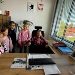 Grupa dzieci stoi w biurze przy biurku z klawiaturą; jedno dziecko siedzi na fotelu. Na ścianie są godła, a przez okno widać budynek z czerwonym dachem. Kolory są jasne i naturalne.