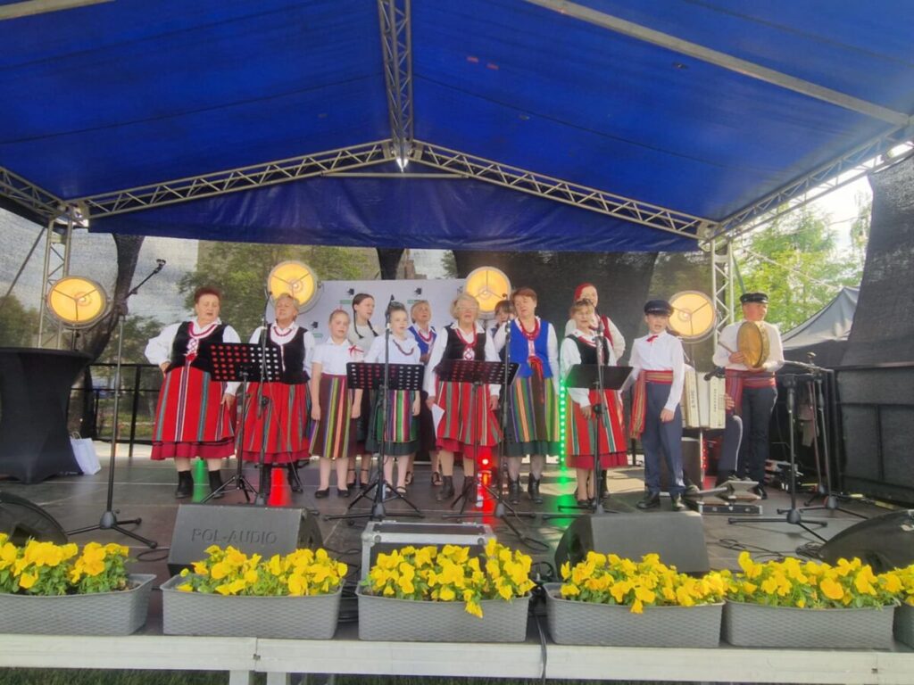 Grupa ludzi w kolorowych strojach ludowych stoi na scenie pod niebieskim zadaszeniem. W tle są drzewa, a mikrofony i światła są umieszczone przed nimi. Na przodzie żółte kwiaty w donicach.