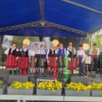 Grupa ludzi w kolorowych strojach ludowych stoi na scenie pod niebieskim zadaszeniem. W tle są drzewa, a mikrofony i światła są umieszczone przed nimi. Na przodzie żółte kwiaty w donicach.