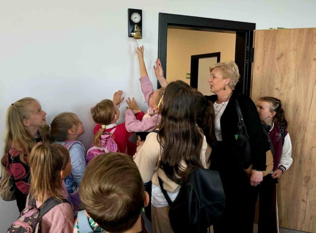 Grupa dzieci i kobieta stoją przy ścianie, gdzie dziecko sięga do dzwonka. Dzieci są kolorowo ubrane z plecakami. Na białej ścianie wisi złoty dzwonek.