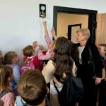 Grupa dzieci i kobieta stoją przy ścianie, gdzie dziecko sięga do dzwonka. Dzieci są kolorowo ubrane z plecakami. Na białej ścianie wisi złoty dzwonek.