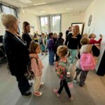 Grupa dzieci z plecakami w jasnej klasie z dużym oknem, otoczona przez dorosłych. W tle biurko i komputer. Blada podłoga, na ścianie obrazek i dzwonek.