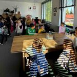 Grupa dzieci w kolorowych strojach i plecakach w klasie. Światło przez żaluzje tworzy pasy na stole. Ściany są jasne, w tle plakaty.