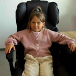 Dziewczynka w różowym swetrze siedzi na czarnym fotelu biurowym. Obok drewniane biurko z przedmiotami. W tle donica z rośliną.