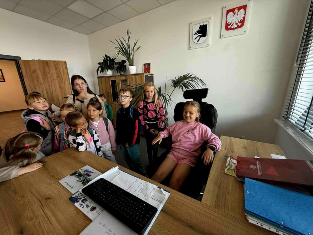 Grupa dzieci stoi w biurze; jedna siedzi za biurkiem na czarnym fotelu. Ściany ozdobione godłami; rośliny w tle. Drewniane biurko z dokumentami i klawiaturą.