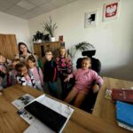 Grupa dzieci stoi w biurze; jedna siedzi za biurkiem na czarnym fotelu. Ściany ozdobione godłami; rośliny w tle. Drewniane biurko z dokumentami i klawiaturą.