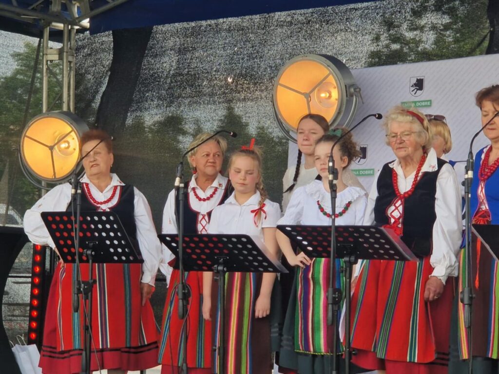 Grupa osób w ludowych strojach występuje na scenie z mikrofonami. Mają kolorowe spódnice i korale. Tło jest ciemne z reflektorami.