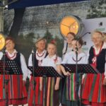 Grupa osób w ludowych strojach występuje na scenie z mikrofonami. Mają kolorowe spódnice i korale. Tło jest ciemne z reflektorami.