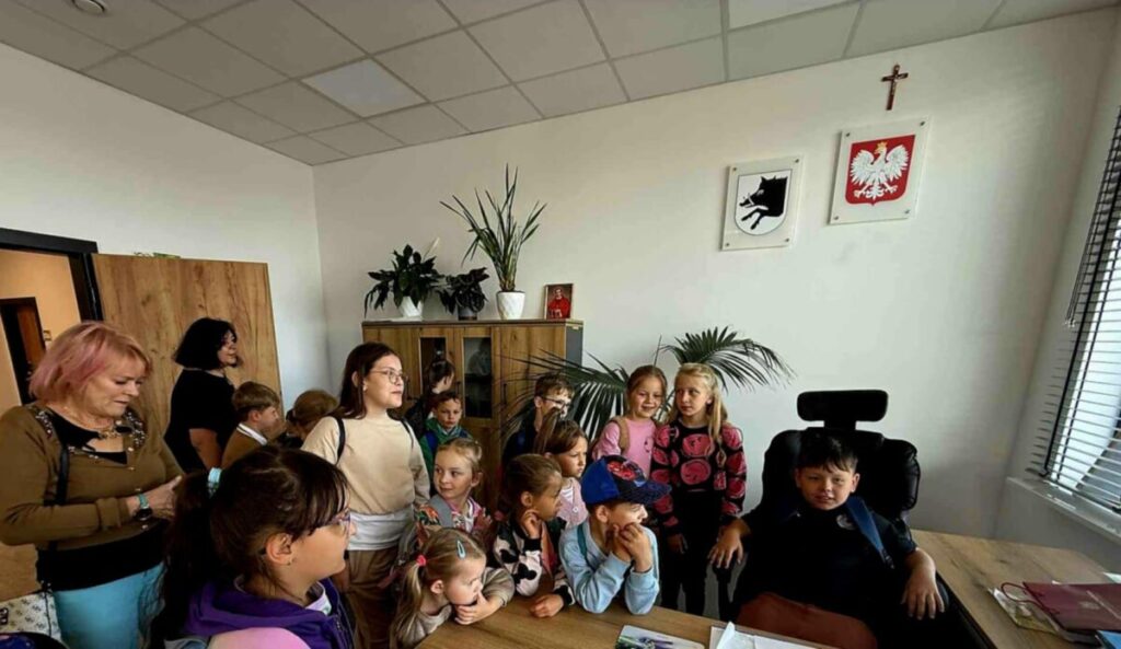 Grupa dzieci w biurze w jasnym pomieszczeniu. Na ścianie wisi godło Polski. W tle rośliny i drewniana szafka.