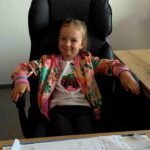 Na zdjęciu dziewczynka siedzi na czarnym fotelu. Ma na sobie kolorową kurtkę i koszulkę z cekinowym wzorem. W tle jest jasna ściana i drewniane biurko z dokumentami.