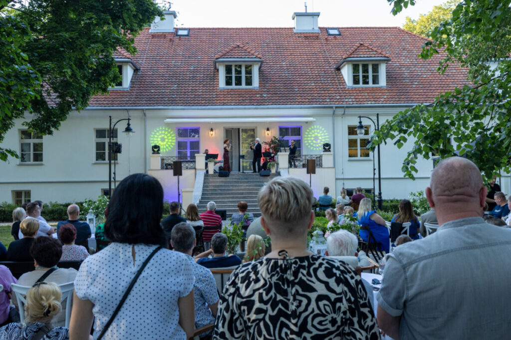 Na zdjęciu widać koncert na tle białego budynku z czerwonym dachem. Przed sceną siedzi publiczność w ogrodzie. W tle widać zielone drzewa.