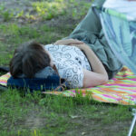 Na zdjęciu osoba leży na kolorowym kocu na trawie, z głową na plecaku. Ma na sobie biały T-shirt z wzorami i zielone spodnie. Tło jest zielone z roślinnością i cieniami.