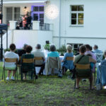 Grupa osób ogląda występ na scenie przed białym budynkiem. Na trawniku siedzą widzowie na krzesłach. Jasne światła dekoracyjne.