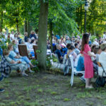 Duża grupa ludzi uczestniczy w wydarzeniu plenerowym w lesie. W tle zielone drzewa, wiele osób siedzi lub stoi. Dominuje zieleń i różnorodne kolory ubrań.