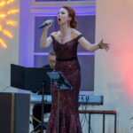 Kobieta w cekinowej sukni śpiewa na scenie, trzymając mikrofon. W tle muzyk przy klawiszach i jasne oświetlenie. Ściana zdobiona wzorem.