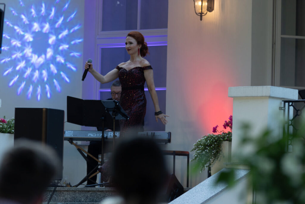 Na scenie stoi kobieta w czerwonej sukni, trzyma mikrofon. Obok niej pianista. Tło oświetlone jest kolorowymi światłami.
