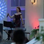 Na scenie stoi kobieta w czerwonej sukni, trzyma mikrofon. Obok niej pianista. Tło oświetlone jest kolorowymi światłami.