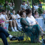 Grupa ludzi siedzi na krzesłach na trawie podczas wydarzenia plenerowego. Tło to drzewa i białe stoły z dekoracjami. Uczestnicy noszą letnie, kolorowe ubrania.