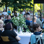 Na zdjęciu widoczna jest duża grupa ludzi siedząca przy stołach na świeżym powietrzu w parku. Na pierwszym planie są białe obrusy i zielone rośliny. Tło wypełniają drzewa i letnia zieleń.