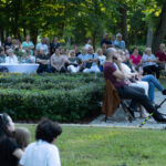 Grupa ludzi siedzi na krzesłach w parku. Tło to bujna zieleń drzew i krzewów. Jasne letnie światło prześwituje przez liście.