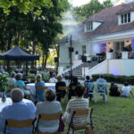 Ogród z koncertem na tle białego budynku. Publiczność siedzi przy stołach, zielone drzewa w tle. Scena jest oświetlona kolorowymi światłami.