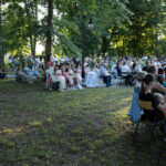 Duża grupa ludzi siedzi na krzesłach na trawie w parku. Dookoła widać drzewa i zielone tło. Przed nimi ustawione są stoły z białymi obrusami.