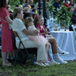 Grupa osób siedzi na krzesłach na trawie podczas plenerowego wydarzenia. Kobiety i dzieci w jasnych ubraniach patrzą na scenę. Obok stoliki z białymi obrusami i roślinami.