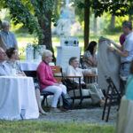 Obraz przedstawia ludzi siedzących przy stołach na otwartej przestrzeni. Starsza kobieta w różowej kurtce siedzi obok mężczyzny z kocem. Tło tworzy zielone, słoneczne otoczenie z drzewami.