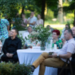Grupa osób siedzi przy stole na zewnątrz, otoczona zielenią. Na stole biały obrus i wazon z kwiatami. Tło to drzewa i trawa.