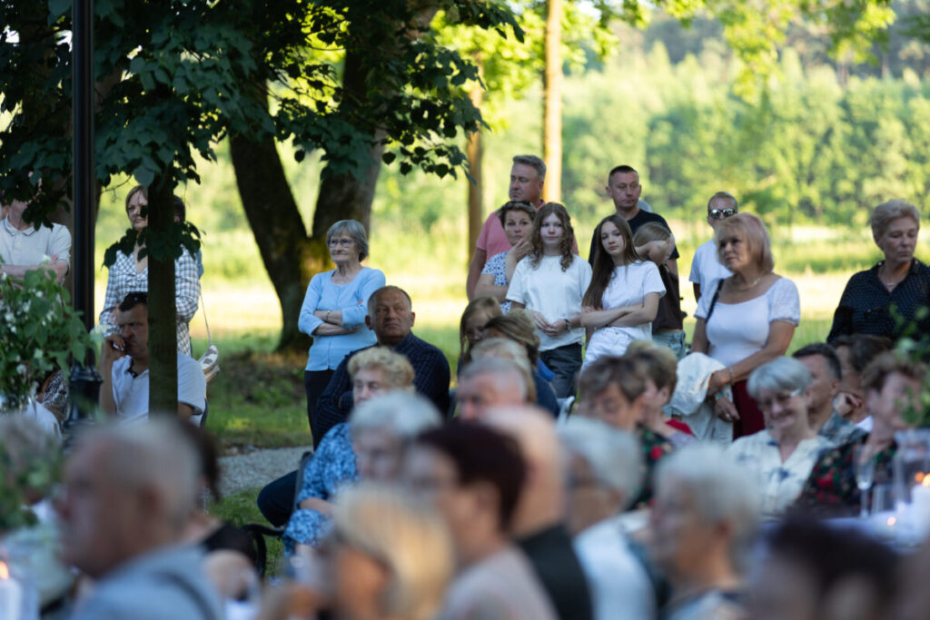 Zdjęcie przedstawia grupę ludzi na świeżym powietrzu, wśród drzew, w letnich ubraniach. W tle widać zieleń i delikatne światło słoneczne. Atmosfera jest pogodna, a ludzie wyglądają na zainteresowanych.