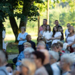 Zdjęcie przedstawia grupę ludzi na świeżym powietrzu, wśród drzew, w letnich ubraniach. W tle widać zieleń i delikatne światło słoneczne. Atmosfera jest pogodna, a ludzie wyglądają na zainteresowanych.