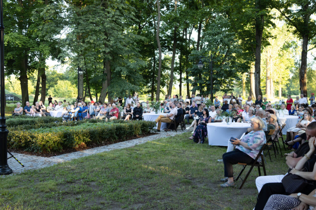 Na zdjęciu widoczna jest grupa ludzi siedzących na krzesłach i przy okrągłych stołach na zewnątrz. Tło stanowi park z zielonymi drzewami. Atmosfera jest spokojna, z naturalnym oświetleniem.