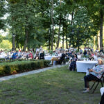 Na zdjęciu widoczna jest grupa ludzi siedzących na krzesłach i przy okrągłych stołach na zewnątrz. Tło stanowi park z zielonymi drzewami. Atmosfera jest spokojna, z naturalnym oświetleniem.