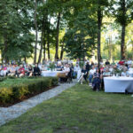 Grupa ludzi siedzi na krzesłach w parku podczas wydarzenia. Stoły przykryte białymi obrusami, zielone drzewa w tle. Słoneczny dzień.