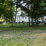 Obraz przedstawia biały budynek wśród drzew, a na trawie stoi grupa ludzi i rowery. Zieleń dominuje w tle. Słoneczne popołudnie.