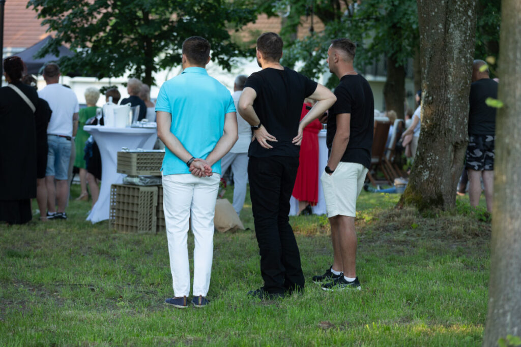 Trzech mężczyzn stoi na trawie, tyłem do kamery. Otoczenie to zielone drzewa i grupa ludzi wokół białych stolików. Jeden mężczyzna ma turkusową koszulkę, reszta jest ubrana w czerń.