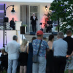 Na zdjęciu widać koncert na świeżym powietrzu. Mężczyzna w czarnym garniturze śpiewa na scenie, przed białym budynkiem. Publiczność siedzi i stoi na trawiastym placu, tło zdobią kolorowe światła.