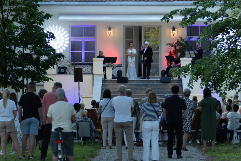 Grupa ludzi ogląda występ na scenie przed eleganckim budynkiem. Światła w tle są kolorowe, a drzewa wokół dodają zieleni. Wykonawcy stoją przy mikrofonach, otoczeni muzykami.