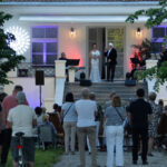 Grupa ludzi ogląda występ na scenie przed eleganckim budynkiem. Światła w tle są kolorowe, a drzewa wokół dodają zieleni. Wykonawcy stoją przy mikrofonach, otoczeni muzykami.