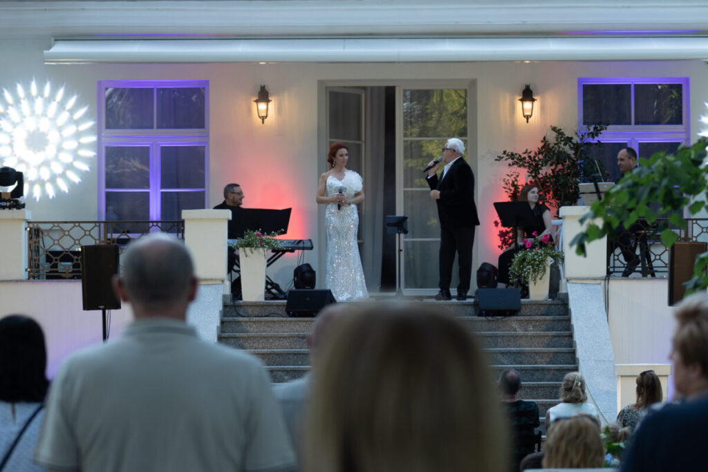 Widok na koncert na zewnątrz, śpiewak w eleganckim stroju na scenie. Publiczność siedzi na schodach. Tło oświetlone, rośliny.