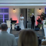 Widok na koncert na zewnątrz, śpiewak w eleganckim stroju na scenie. Publiczność siedzi na schodach. Tło oświetlone, rośliny.