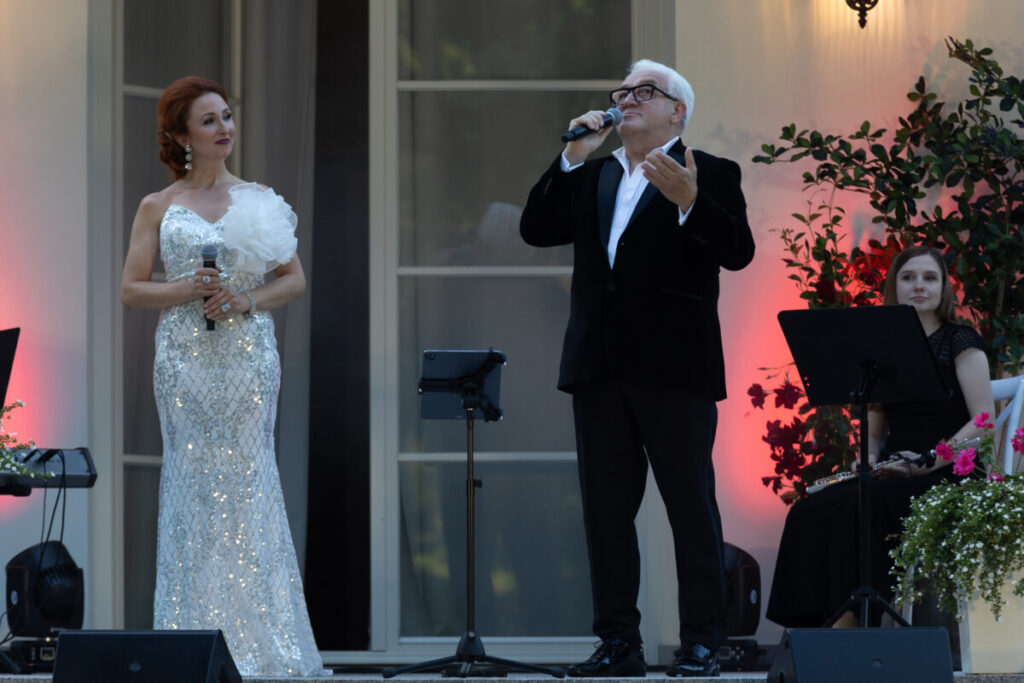 Dwóch artystów występuje na scenie, ubrani elegancko. Kobieta w białej, błyszczącej sukni, mężczyzna w ciemnym garniturze. Tło z roślinami i ciepłym oświetleniem.