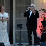 Dwóch artystów występuje na scenie, ubrani elegancko. Kobieta w białej, błyszczącej sukni, mężczyzna w ciemnym garniturze. Tło z roślinami i ciepłym oświetleniem.