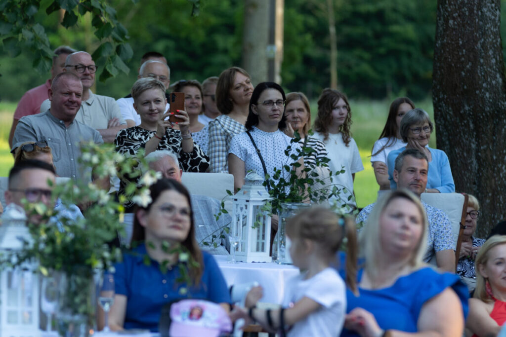 Grupa ludzi na zewnątrz, siedzących przy stołach pod drzewami. Tło jest zielone i naturalne. Dekoracje w bieli i zieleni.