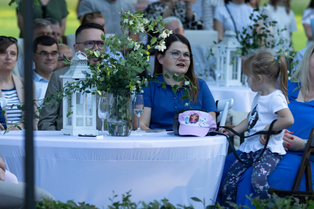 Na zdjęciu grupa ludzi przy strojnie udekorowanym stole na świeżym powietrzu. Na pierwszym planie biała latarnia i bukiet kwiatów. Dziewczynka w jasnej czapce i koszulce siedzi obok kobiety w niebieskiej sukience.