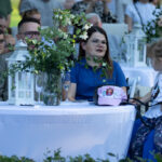 Na zdjęciu grupa ludzi przy strojnie udekorowanym stole na świeżym powietrzu. Na pierwszym planie biała latarnia i bukiet kwiatów. Dziewczynka w jasnej czapce i koszulce siedzi obok kobiety w niebieskiej sukience.