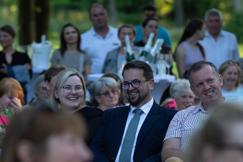 Grupa ludzi siedzi na zewnątrz, większość się uśmiecha. W tle zieleń i stół z lodem i napojami. Atmosfera jest radosna.