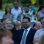 Grupa ludzi siedzi na zewnątrz, większość się uśmiecha. W tle zieleń i stół z lodem i napojami. Atmosfera jest radosna.