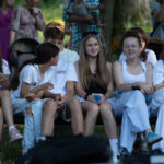 Grupa młodych osób siedzi na ławce, ubrane w jasne stroje. W tle widać ludzi w parku, drzewa i zieleń. Jest pogodny dzień, niektórzy trzymają telefony.