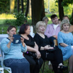 Grupa osób siedzi na krzesłach na świeżym powietrzu. Tło to zielone drzewa i trawnik. Stół pokryty białym obrusem, na nim kieliszki.